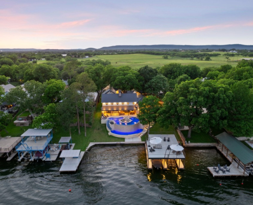 Wide drone view of lakefront home, pool, and Hill Country countryside at dusk